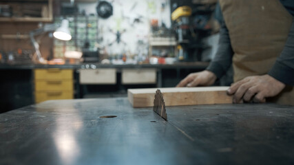 A carpenter wearing an apron and a mask is sawing a tree on a circular saw in a workshop