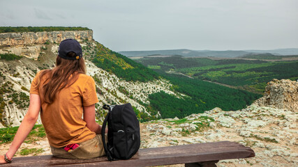 A girl tourist sits on a wooden bench on the top of the Crimean mountains, which offers a beautiful view. © Michael