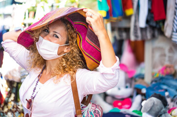 A smiling attractive woman with blonde and curly hair wearing a medical mask due to coronavirus standing at the flea market trying on a beautiful multi colored hat
