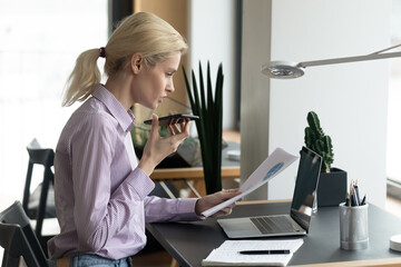 Concentrated smart young blonde businesswoman reading paper marketing research report, holding...