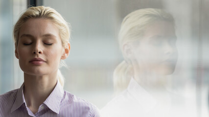 Head shot close up mindful young businesswoman breathing fresh air with closed eyes near window,...