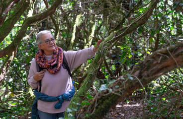 Active senior woman white haired enjoying mountain hike in nature in the middle of the forest with backpack on shoulders
