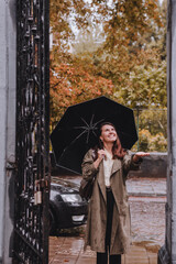 young pretty woman in autumn outfit walking by city street with umbrella at rainy weather