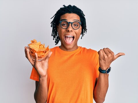 Young african american man holding nachos potato chips pointing thumb up to the side smiling happy with open mouth