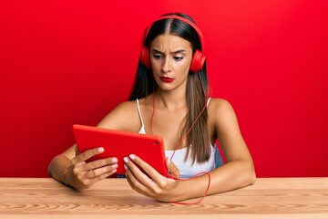 Young hispanic woman sitting on the table using touchpad wearing headphones clueless and confused...