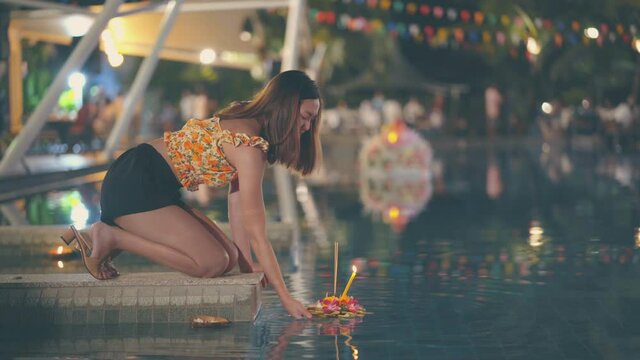 The woman is floating banana leaf krathong at the pool in Loy Krathong festival with lightings and flags decorated around the pool 4K.