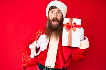 Handsome young red head man with long beard wearing santa claus costume and big bag with presents clueless and confused expression. doubt concept.