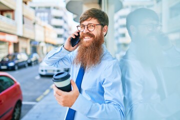 Young handsome redhead businessman smiling happy. Leaning on the wall with smile on face using smartphone drinking take away coffee at street of city.