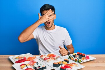 Young handsome man eating sushi sitting on the table peeking in shock covering face and eyes with hand, looking through fingers with embarrassed expression.