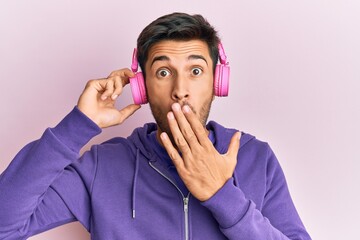 Young handsome man listening to music using headphones covering mouth with hand, shocked and afraid...