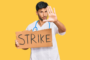 Young handsome man wearing doctor uniform holding strike banner cardboard with open hand doing stop sign with serious and confident expression, defense gesture
