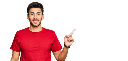 Young handsome man wearing casual red tshirt with a big smile on face, pointing with hand finger to the side looking at the camera.