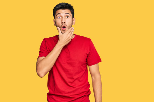 Young handsome man wearing casual red tshirt looking fascinated with disbelief, surprise and amazed expression with hands on chin