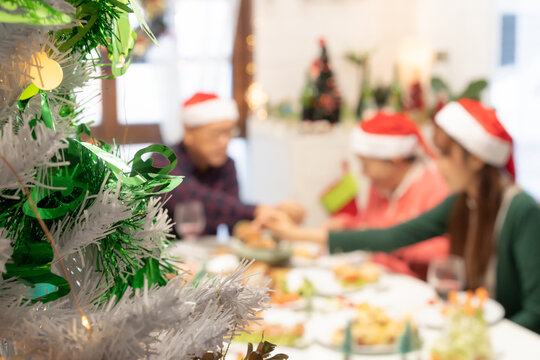 Close-up of half decorated Christmas tree on blurred image of happy Asian family with Santa Claus hats eating Christmas dinner together and grandfather serving food to grandmother.