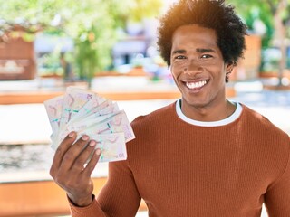 Young african american man smiling happy. Standing with smile on face holding colombian pesos banknotes at town street.