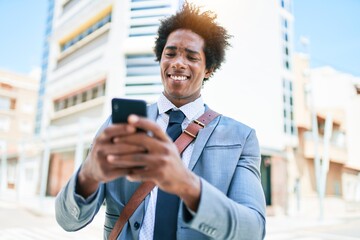 Young african american businessman wearing suit smiling happy. Standing with smile on face using smartphone at town street.