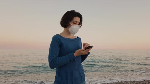 Ragazza con mascherina parla al telefono in spiaggia vicino al mare