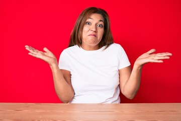 Middle age brunette hispanic woman wearing casual style sitting on the table clueless and confused...
