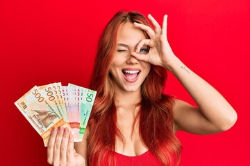 Young beautiful redhead woman holding norwegian krone banknotes smiling happy doing ok sign with...