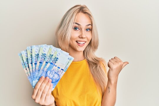 Beautiful caucasian blonde girl holding south african 100 rand banknotes pointing thumb up to the side smiling happy with open mouth