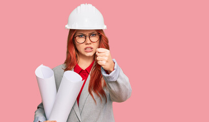 Young redhead architect woman wearing safety helmet holding blueprints annoyed and frustrated shouting with anger, yelling crazy with anger and hand raised