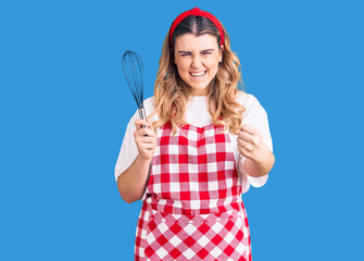 Young caucasian woman wearing apron holding whisk annoyed and frustrated shouting with anger, yelling crazy with anger and hand raised