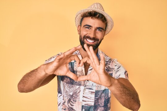 Young hispanic man wearing summer hat smiling in love doing heart symbol shape with hands. romantic concept.