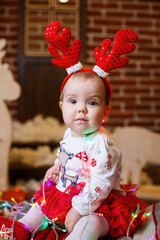 A little girl in warm sweater sits under a Christmas tree with toys and gifts with horns on her head. Happy childhood. New Year holiday atmosphere