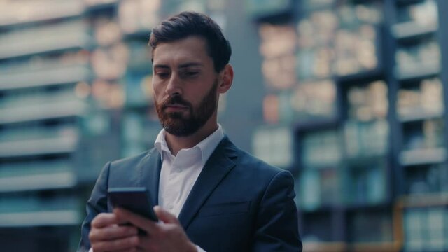 Adult successful caucasian serious businessman in formal outfit walking in corporate business district with phone browsing financial news smartphone application.