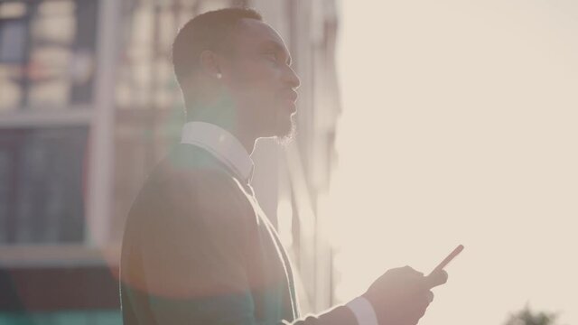 Young attractive businessman with phone at beautiful morning sunlight. Afro-american confident entrepreneur browsing smartphone applications standing outdoor.