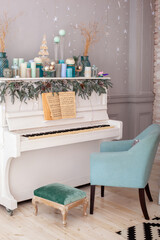 Interior living room is decorated in classic style with white piano, Christmas tree and gifts. New Year decoration. Piano in room decorated candles for Christmas. Piano with notes and blue armchair	