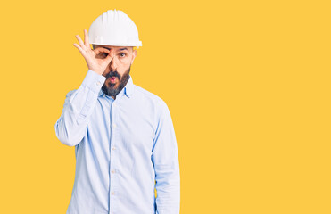 Young handsome man wearing architect hardhat doing ok gesture shocked with surprised face, eye looking through fingers. unbelieving expression.