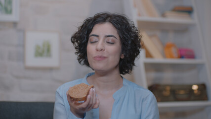 A cheerful young woman is sitting at home on an armchair and eating peanut butter bread.