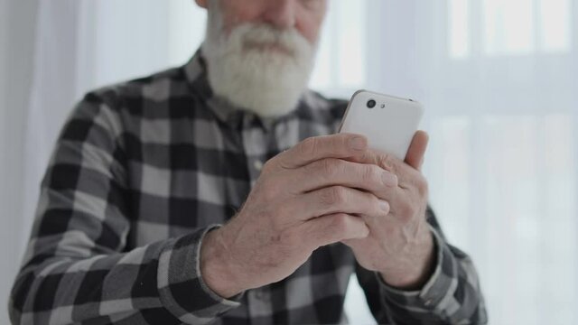 Positive senior man typing on smartphone indoor, chatting or looking at photos