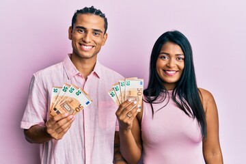 Young latin couple holding 50 euro banknotes looking positive and happy standing and smiling with a...