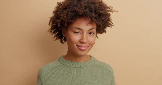 Close up of beautiful mysterious young Afro American woman looks directly at camera winks eye to someone smiles pleasantly being in good mood wears casual clothes isolated over brown background