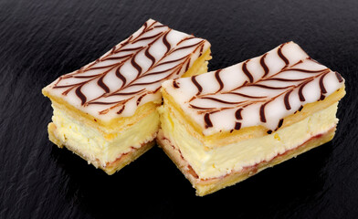 Fresh dairy cream filled slices made with puff pastry topped with icing and chocolate flavour decoration on a slate stone background