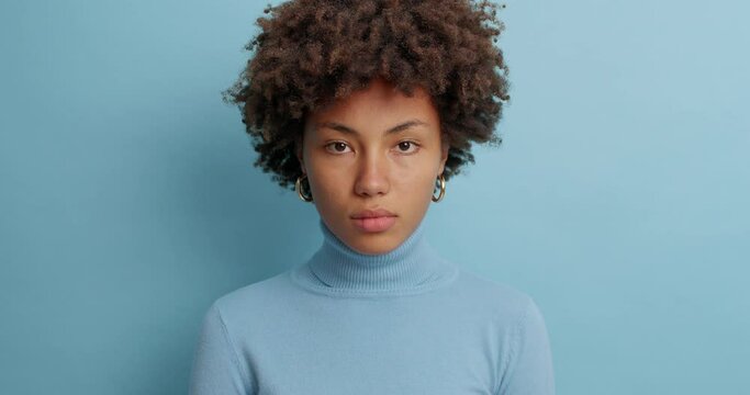 Attractive serious woman with curly hairstyle looks directly at camera dressed in casual turtleneck winks eyes has calm face expression isolated on blue wall. Assertive young Afro American female