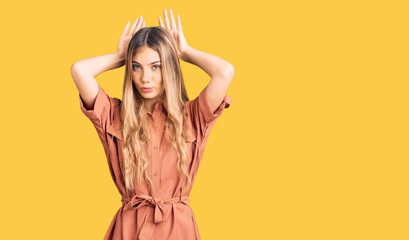 Beautiful caucasian woman with blonde hair wearing summer jumpsuit doing bunny ears gesture with...