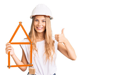 Beautiful caucasian woman with blonde hair wearing architect hardhat and holding tools smiling happy and positive, thumb up doing excellent and approval sign