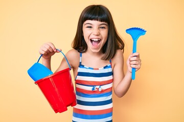Young little girl with bang playing with summer shovel and bucket toys smiling and laughing hard out loud because funny crazy joke. © Krakenimages.com