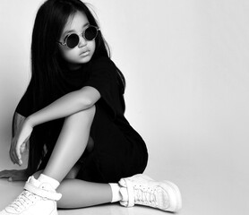 Portrait of brunette asian girl wearing stylish black dress, sneakers and sunglasses sitting on floor and looking aside