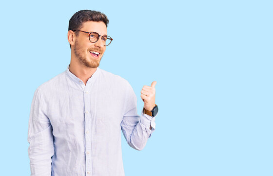 Handsome young man with bear wearing elegant business shirt and glasses smiling with happy face looking and pointing to the side with thumb up.