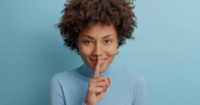 Mysterious dark skinned woman keeps index finger on lips tells secret makes silence gesture stands quiet indoor has curious happy look at camera dressed in turtleneck isolated over blue background