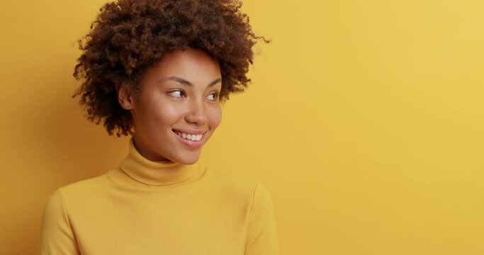 Pleased smiling woman with natural curly hair has beaming smile turns away and grins as sees awesome advertisement dressed in casual turtleneck isolated over yellow background. Happy feelings