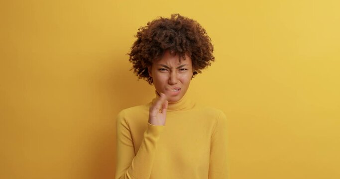 Unhappy dissatisfied dark skinned woman waves palm near nose smells unpleasant stench dressed in casual wear poses over yellow background sees something disgusting. Nasty feelings. Avoiding bad smell
