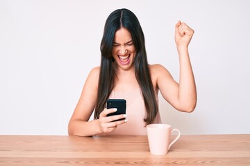 Young caucasian woman sitting at the desk using smartphone drinking coffee screaming proud, celebrating victory and success very excited with raised arms