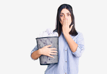 Young beautiful girl holding paper bin full of crumpled papers covering mouth with hand, shocked and afraid for mistake. surprised expression