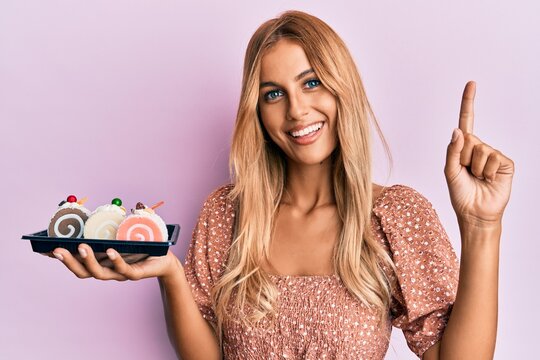 Beautiful blonde young woman holding cake sweets smiling with an idea or question pointing finger with happy face, number one