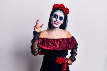 Young woman wearing mexican day of the dead makeup smiling and confident gesturing with hand doing small size sign with fingers looking and the camera. measure concept.